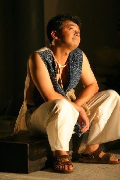 Stage still from He-Me-She-It with a performer seated on a red floor.