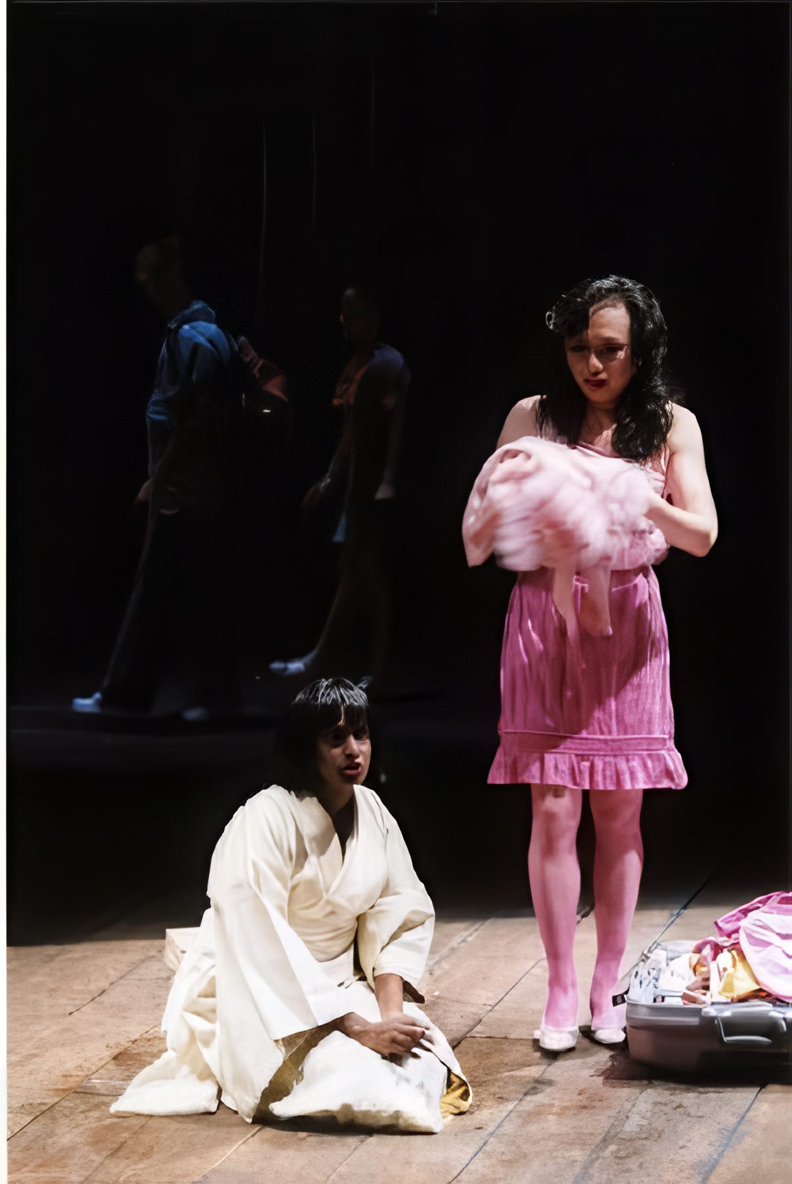 Two performers in a pool of light on a bare wooden stage — one kneeling in a white robe, one standing in pink holding a bundle — others in shadow behind.