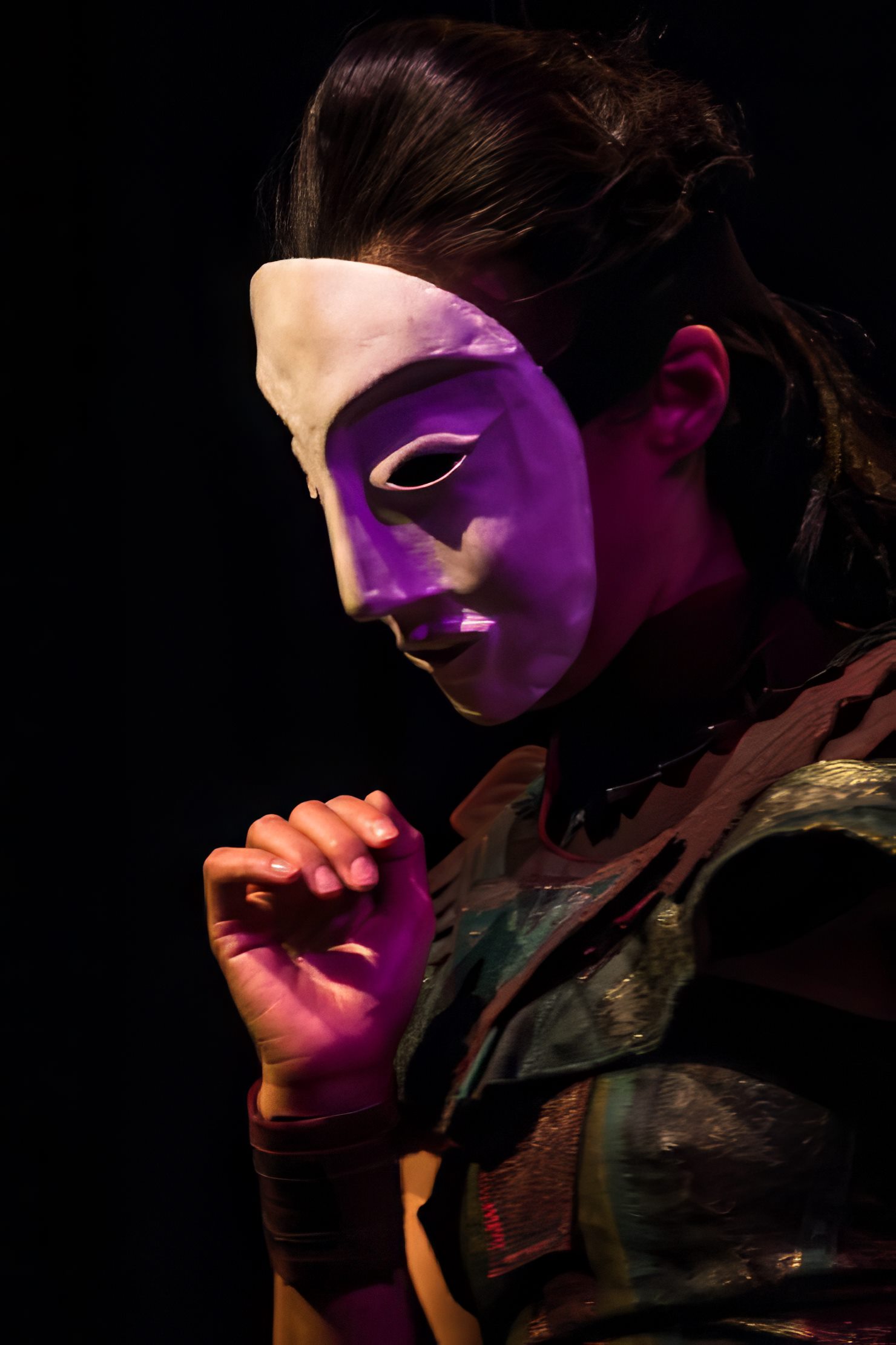 A performer looks downward in a bone-white mask washed in pale violet light, one hand raised, against black.