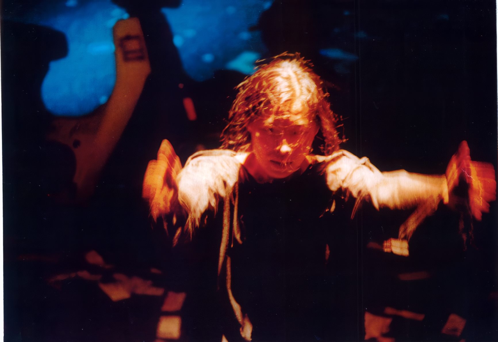 Narumol Thammapruksa mid-performance, arms spread wide, lit amber against a deep blue projection.