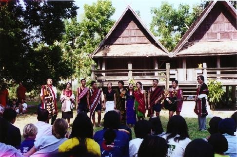 Singers in woven textiles perform in a line before Lanna teak houses, audience seated on the grass.